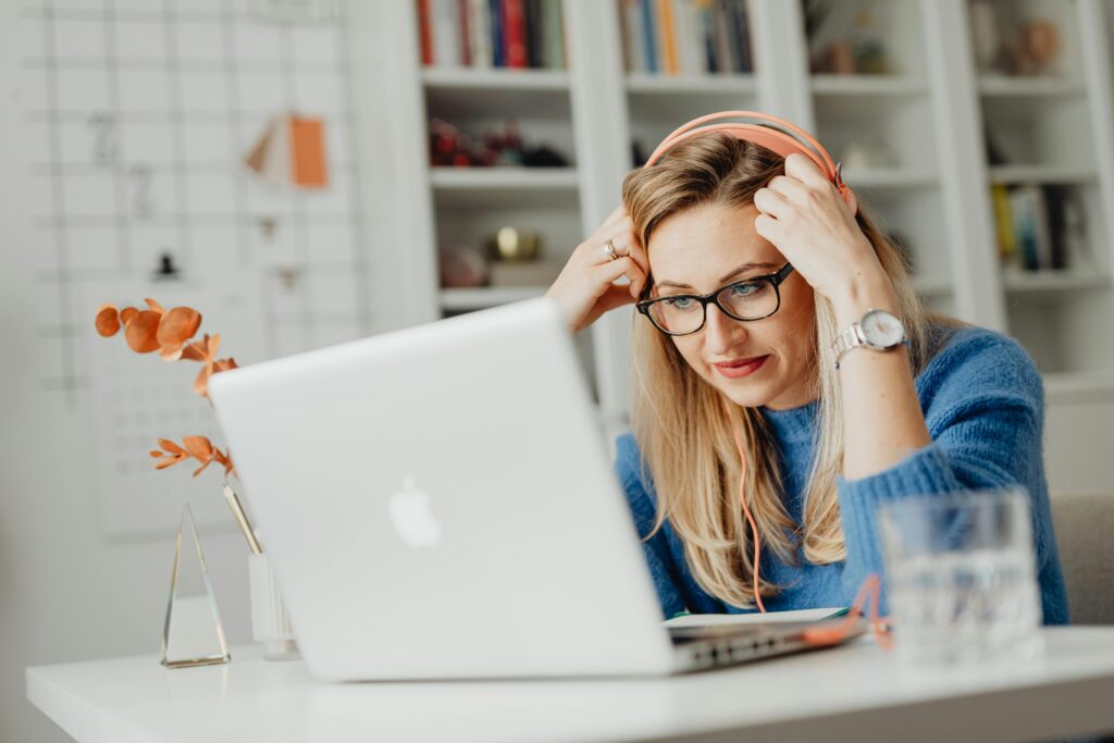 pexels-photo-5908764-5908764 Blond woman wearing eyeglasses and headphones, working on a laptop indoors at a desk.