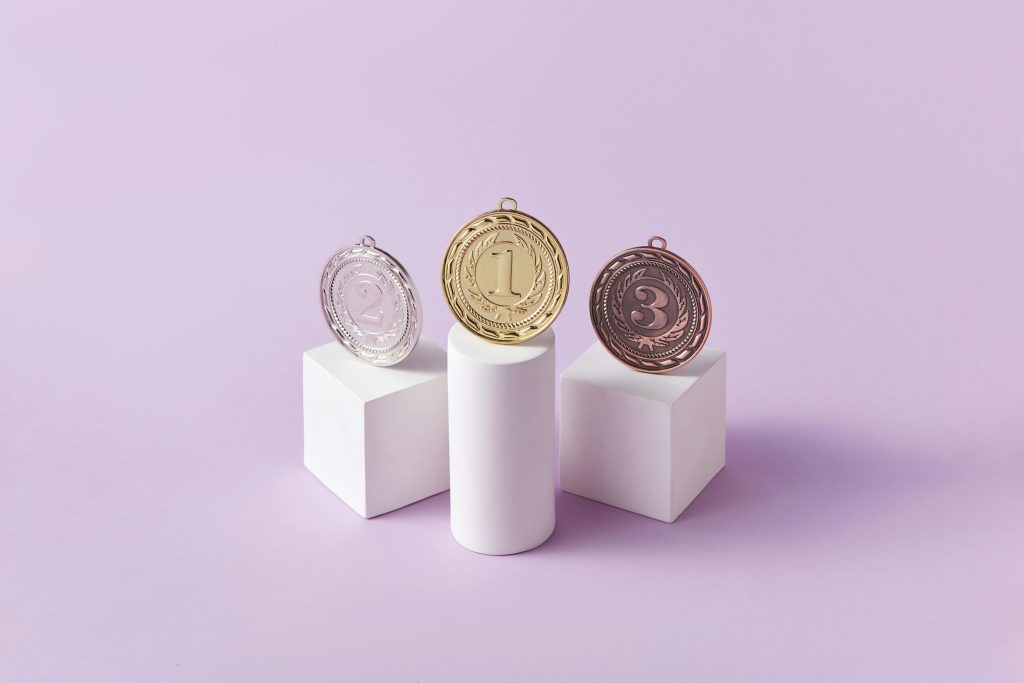 Gold, silver, and bronze medals displayed on white pedestals with a pink background.