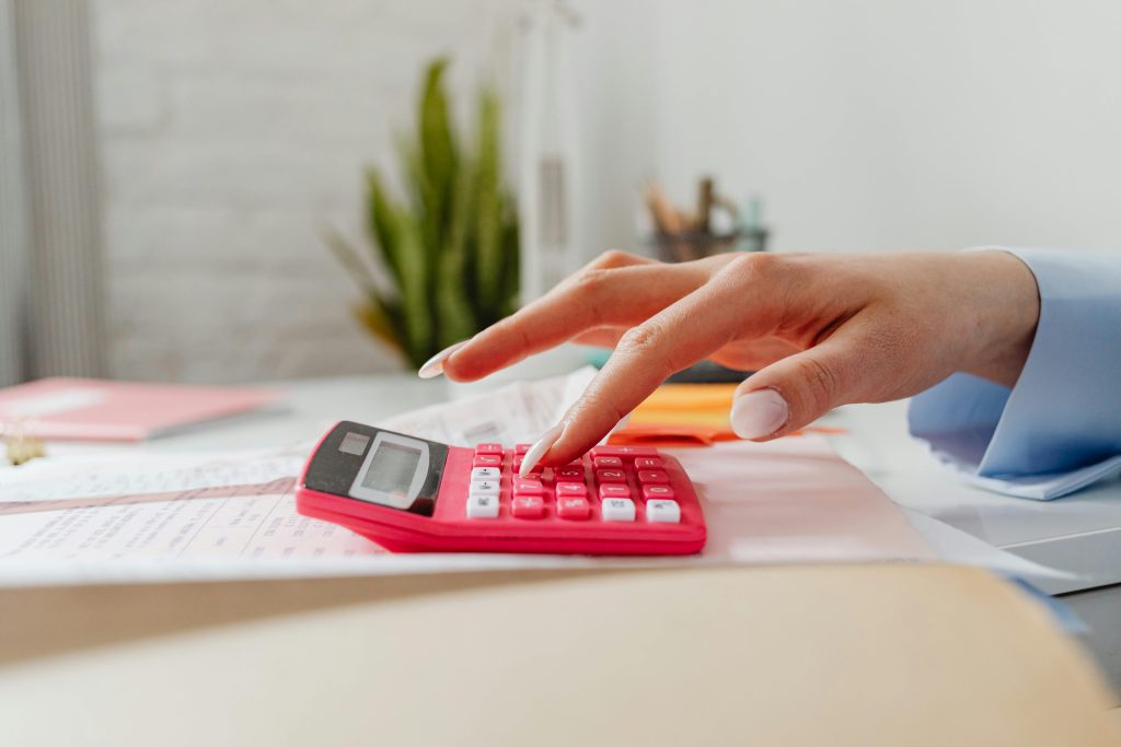 A close-up image of a person's hand using a red calculator on a desk with paperwork.